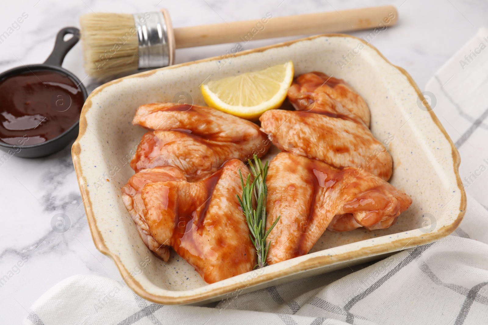 Photo of Fresh marinade, basting brush, raw chicken wings, rosemary and lemon on white marble table, closeup