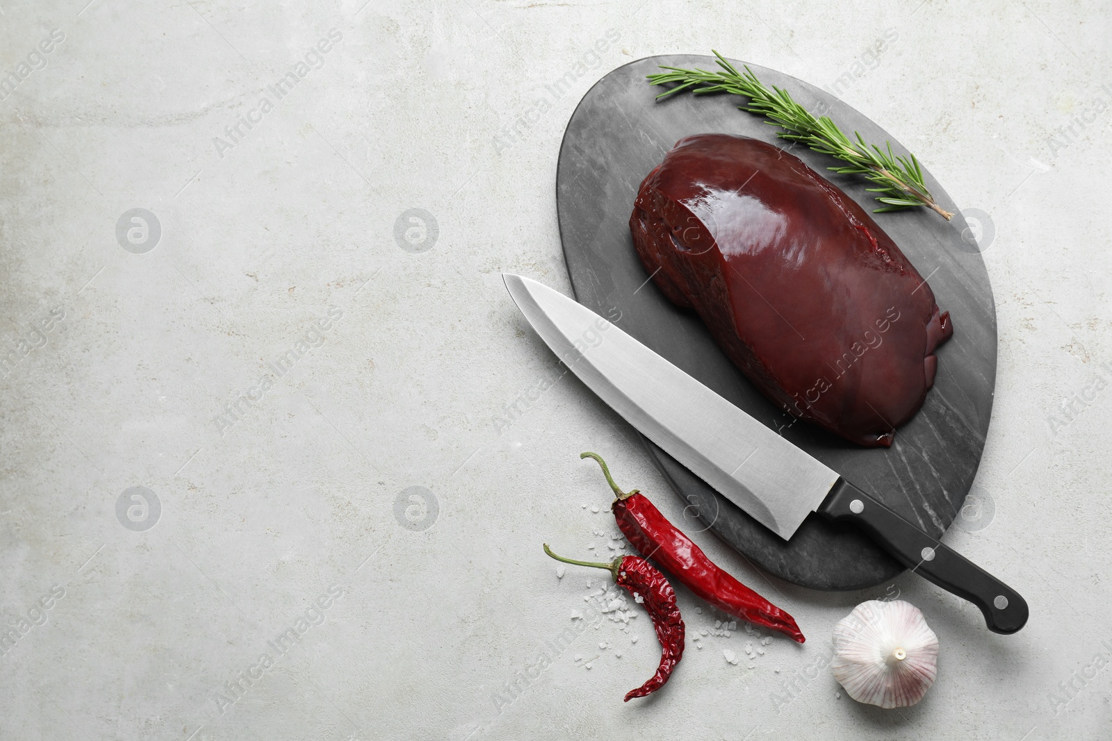 Photo of Piece of raw beef liver with chili peppers, rosemary, garlic and knife on light table, flat lay. Space for text