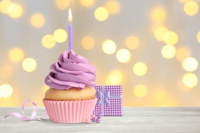 Image of Delicious birthday cupcake with candle and gift box on white wooden table against blurred lights. Space for text