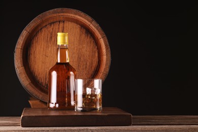 Whiskey with ice cubes in glass, bottle and barrel on wooden table against black background, space for text