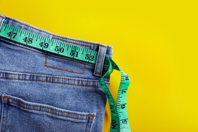 Jeans with measuring tape on yellow background, top view and space for text. Weight loss concept