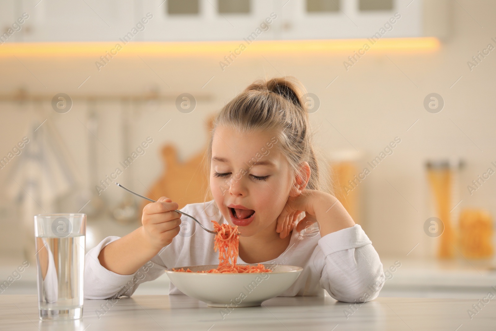 Photo of Cute little girl eating tasty pasta at table in kitchen