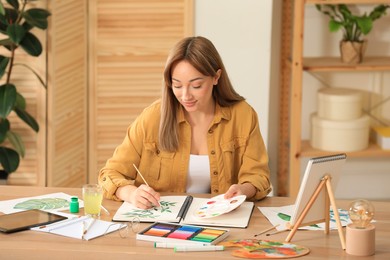 Photo of Young woman painting green twig in sketchbook at wooden table indoors