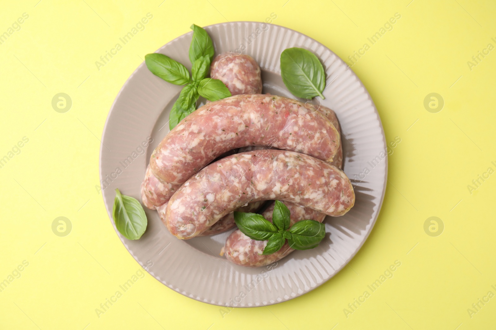 Photo of Raw homemade sausages and basil leaves on yellow background, top view