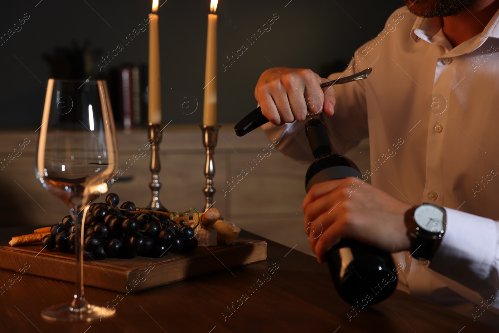 Photo of Romantic dinner. Man opening wine bottle with corkscrew at table indoors, closeup