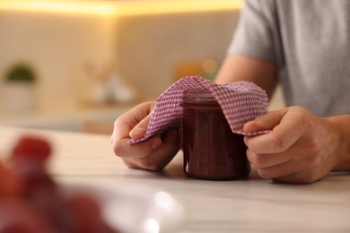 Man packing jar of jam into beeswax food wrap at light table in kitchen, closeup