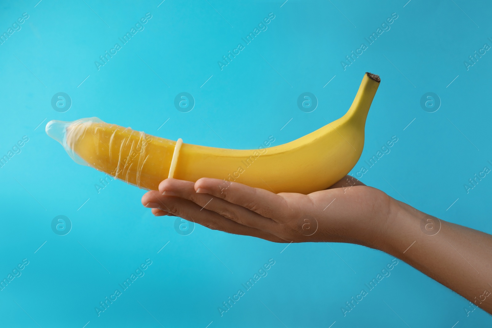 Photo of Woman holding banana in condom on light blue background, closeup. Safe sex concept