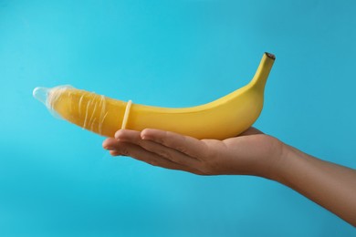 Photo of Woman holding banana in condom on light blue background, closeup. Safe sex concept