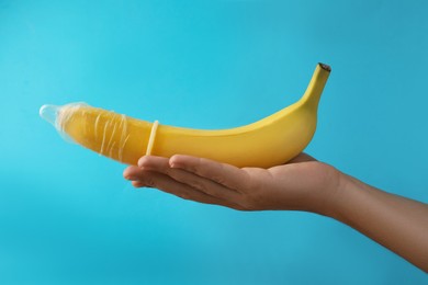 Woman holding banana in condom on light blue background, closeup. Safe sex concept