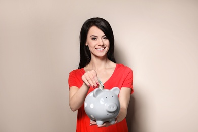 Beautiful woman putting money into piggy bank on color background