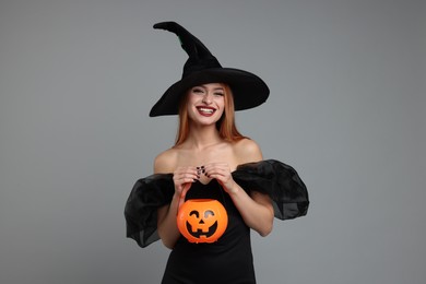 Photo of Happy young woman in scary witch costume with pumpkin bucket on light grey background. Halloween celebration