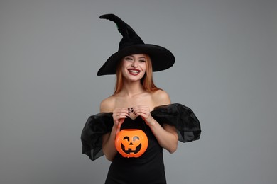 Happy young woman in scary witch costume with pumpkin bucket on light grey background. Halloween celebration