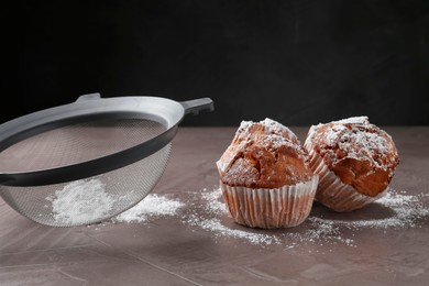 Sieve with sugar powder and muffins on grey textured table, closeup