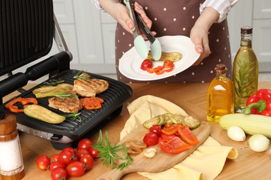 Woman cooking different products with electric grill at wooden table in kitchen, closeup