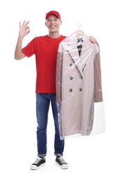 Photo of Dry-cleaning delivery. Happy courier holding coat in plastic bag and showing OK gesture on white background