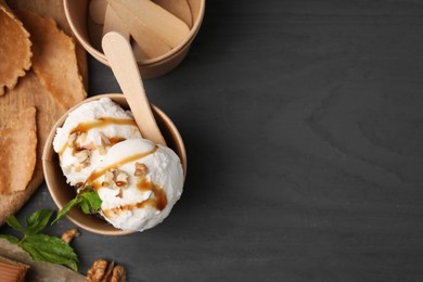 Photo of Tasty ice cream with caramel sauce, mint and nuts in paper cup on grey wooden table, flat lay. Space for text