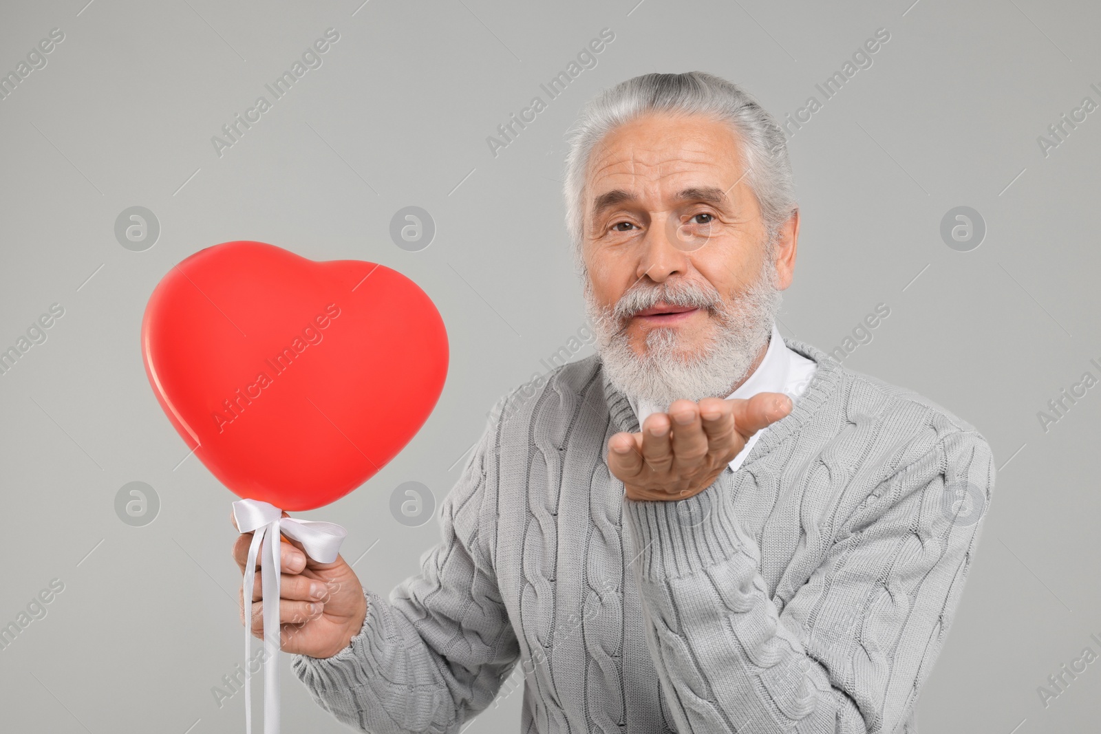 Photo of Senior man with red heart shaped balloon blowing kiss on light grey background