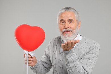 Senior man with red heart shaped balloon blowing kiss on light grey background