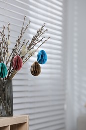 Beautiful pussy willow branches with paper eggs in vase on shelving unit at home, space for text. Easter decor