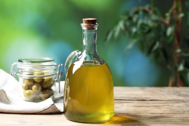 Jug of cooking oil and jar with olives on wooden table against blurred background. Space for text