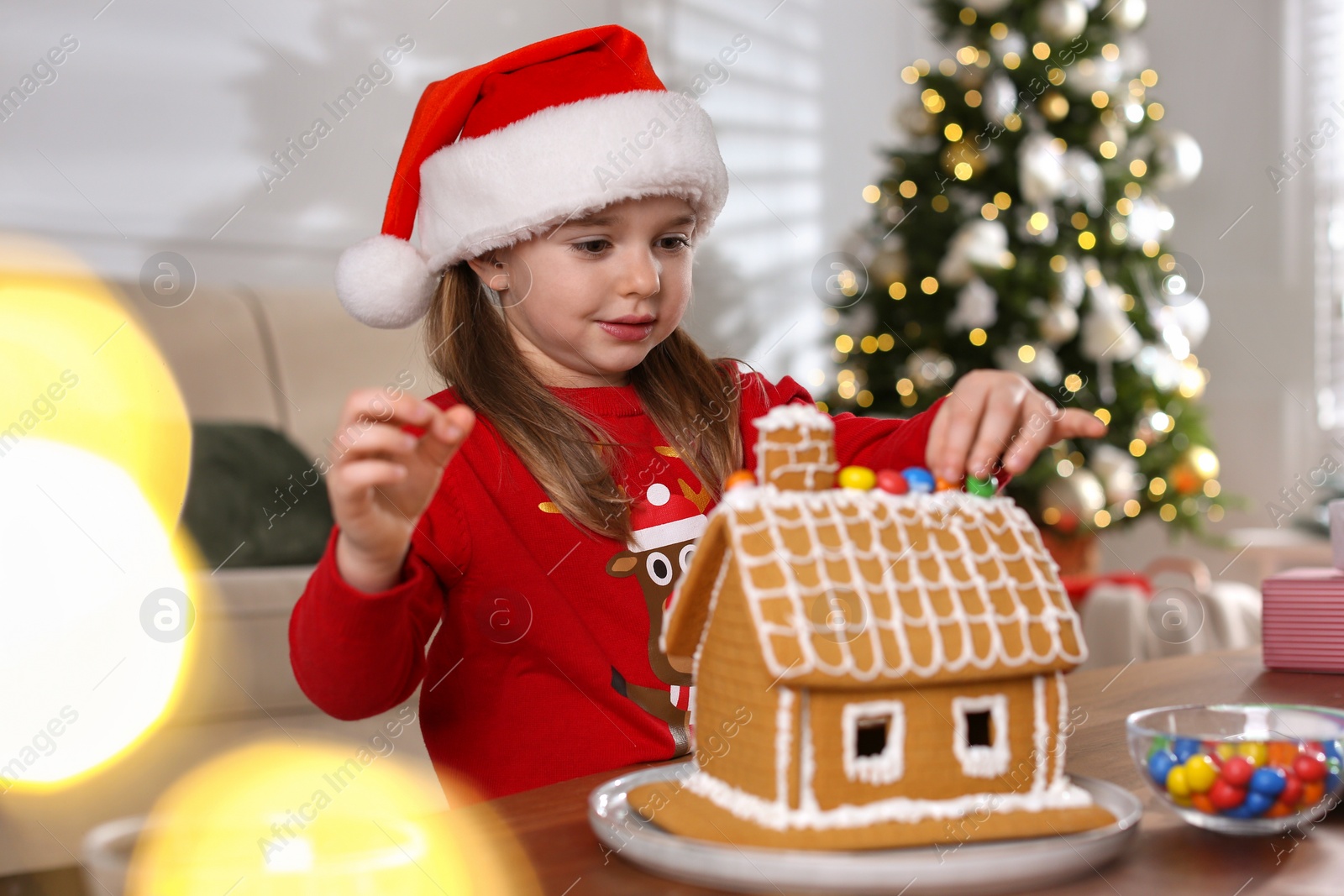 Photo of Cute little girl decorating gingerbread house at table indoors