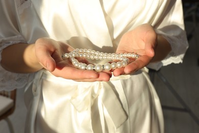 Photo of Bride holding beautiful pearl necklace indoors, closeup. Wedding day