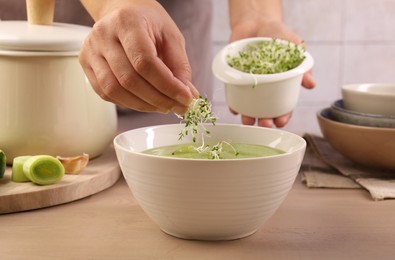 Photo of Woman adding fresh microgreens into bowl with tasty leek soup at wooden table, closeup