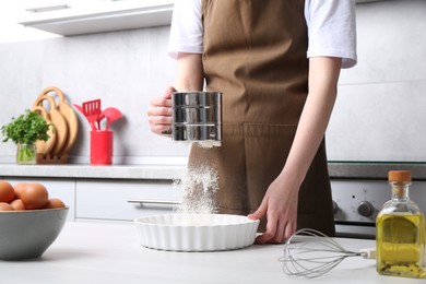 Photo of Woman sieving flour into baking dish at table in kitchen, closeup