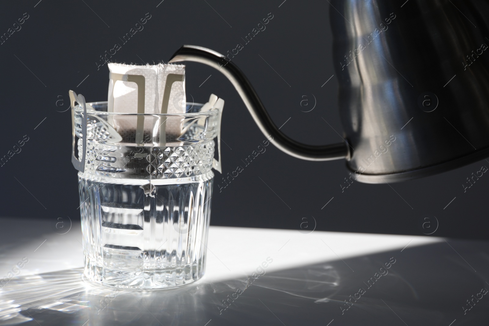 Photo of Glass with drip coffee bag and metal kettle on light grey table, closeup