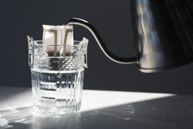 Glass with drip coffee bag and metal kettle on light grey table, closeup