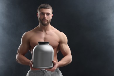 Young man with muscular body holding jar of protein powder on dark grey background, space for text