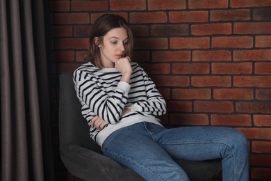 Photo of Sad young woman sitting on chair near brick wall indoors