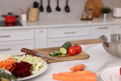 Photo of Cooking vegetarian meal. Fresh vegetables, knife, board and bowl on white marble table in kitchen