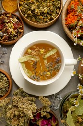 Photo of Cup of aromatic freshly brewed tea surrounded by different dry herbs on grey table, flat lay