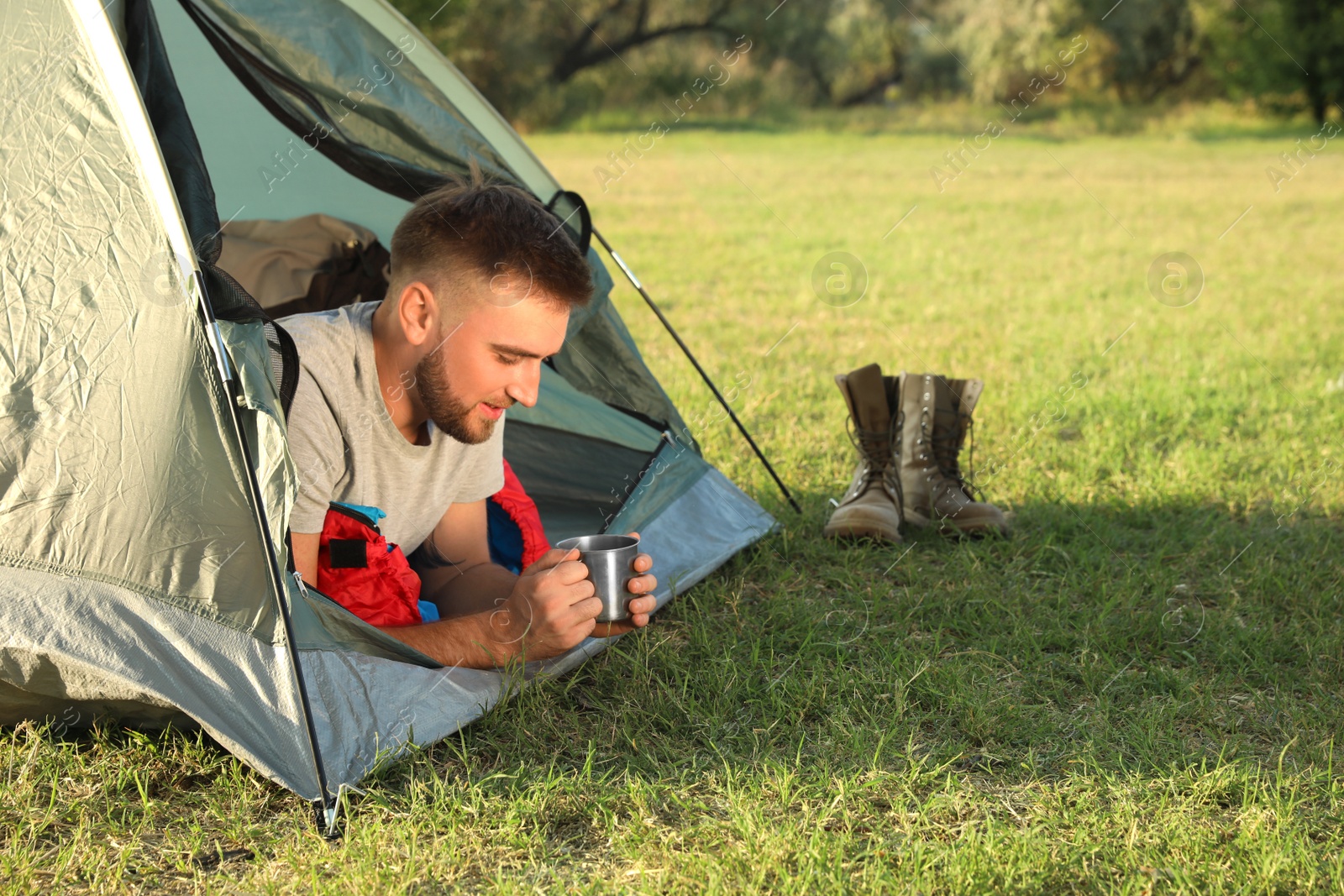 Photo of Young man in sleeping bag with cup of drink lying inside camping tent