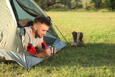 Young man in sleeping bag with cup of drink lying inside camping tent