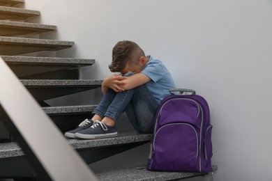 Upset little boy with backpack sitting on stairs indoors