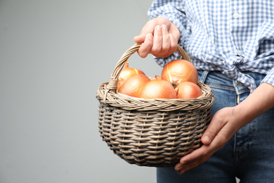 Woman holding wicker basket with raw yellow onion bulbs on grey background, closeup