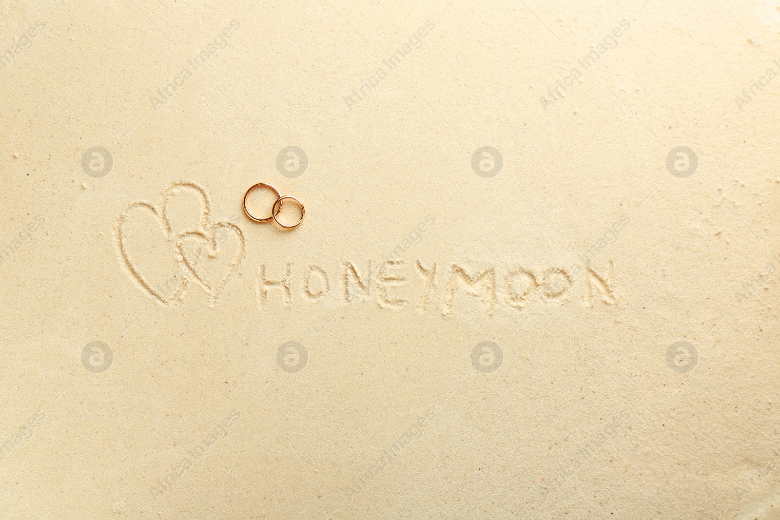 Photo of Word Honeymoon written on sand, hearts two golden rings, top view