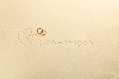 Photo of Word Honeymoon written on sand, hearts two golden rings, top view