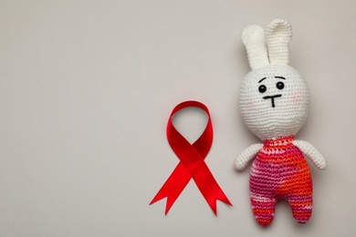 Photo of Cute knitted toy bunny and red ribbon on beige background, flat lay with space for text. AIDS disease awareness