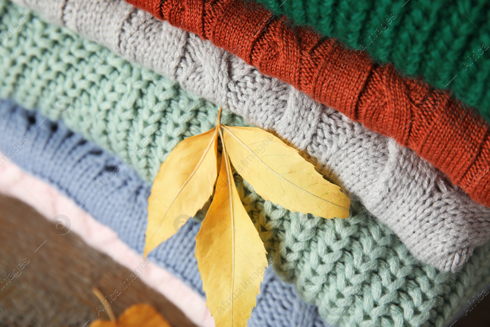 Photo of Stack of warm knitted clothes and autumn leaves on wooden background, closeup