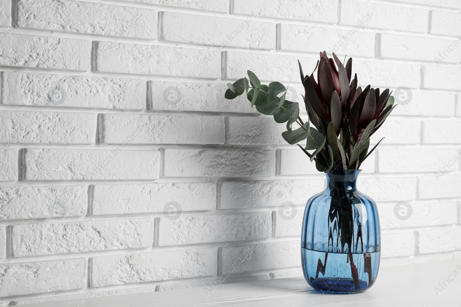 Photo of Bouquet of beautiful protea flowers and eucalyptus branches in glass vase on white wooden table near brick wall. Space for text