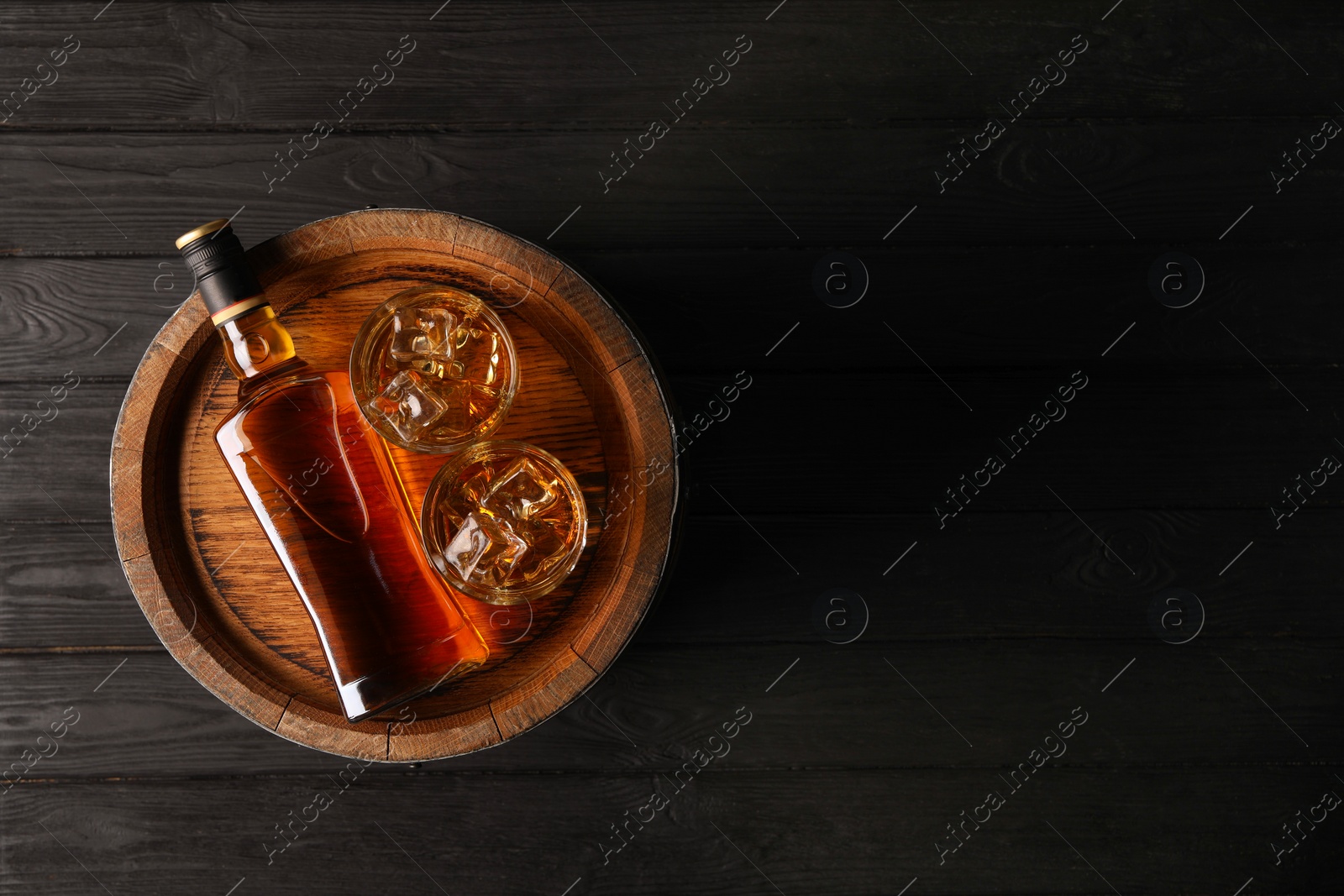 Photo of Whiskey with ice cubes in glasses, bottle and barrel on black wooden table, top view. Space for text