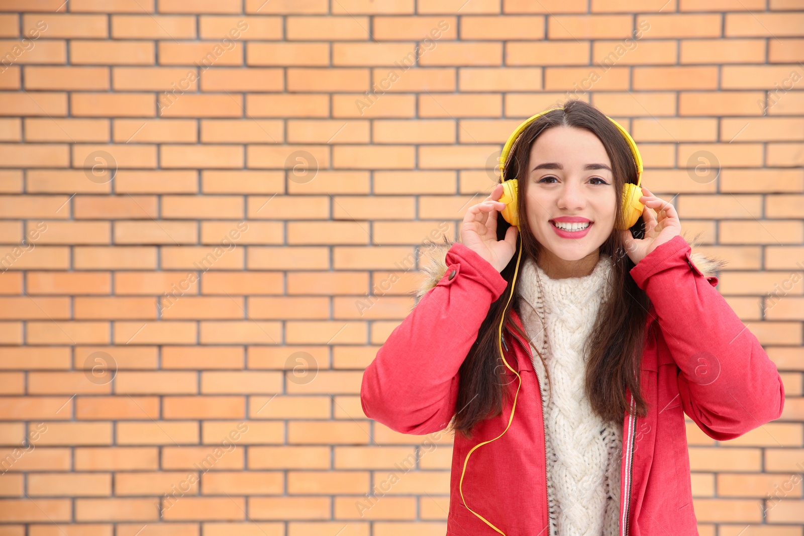 Photo of Beautiful young woman listening to music with headphones against brick wall. Space for text