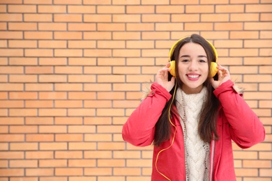 Beautiful young woman listening to music with headphones against brick wall. Space for text