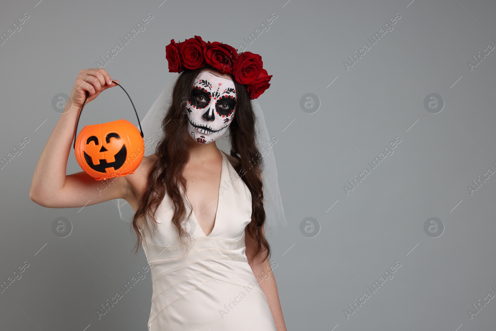 Photo of Young woman in scary bride costume with sugar skull makeup, flower crown and pumpkin bucket on light grey background, space for text. Halloween celebration