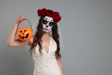 Photo of Young woman in scary bride costume with sugar skull makeup, flower crown and pumpkin bucket on light grey background, space for text. Halloween celebration