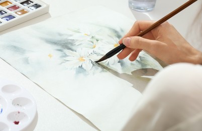 Photo of Woman painting flowers with watercolor at white table, closeup. Creative artwork