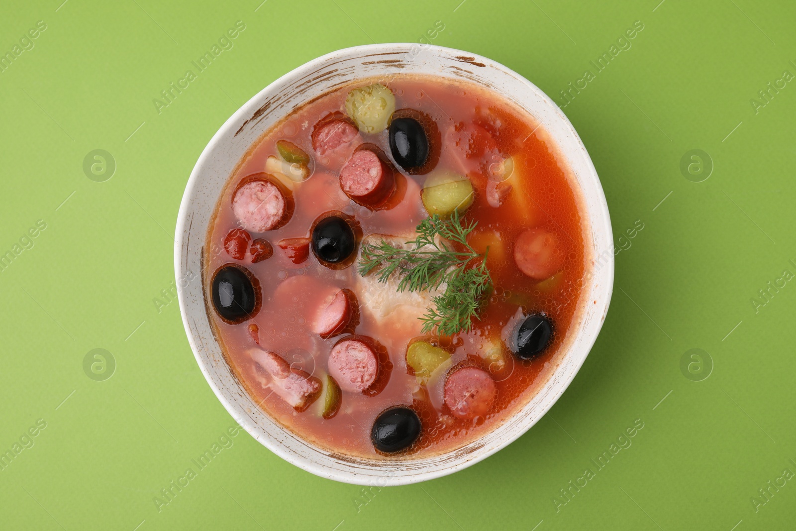 Photo of Meat solyanka soup with thin dry smoked sausages in bowl on light green background, top view